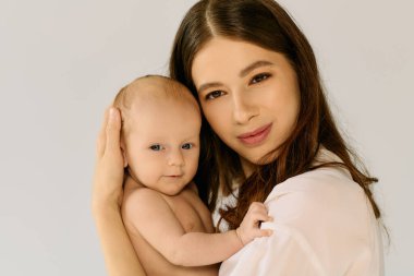 Genç bir anne, gülümseyen bebeğini nazikçe kucağına alır. Sıcak bir ortamda sıcaklık ve şefkat saçar..