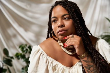 Saçları örgülü, kendinden emin bir Afrikalı Amerikalı kadın yeşil bitkilerin arasında yaz elbisesi içinde poz veriyor..