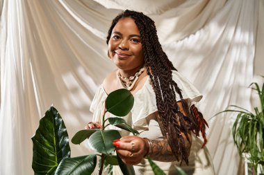 Saçları örgülü neşeli bir kadın, parlak yeşil bitkilerle çevrili açık yaz elbisesi içinde poz veriyor..