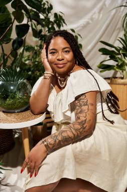 Yazlık elbiseli afro-amerikan bir kadın canlı yeşil bitkilerin arasında poz veriyor, neşe saçıyor..