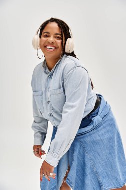 Güzel örgülü bir kadın şık bir stüdyoda neşe içinde poz verirken kot elbise giyer..