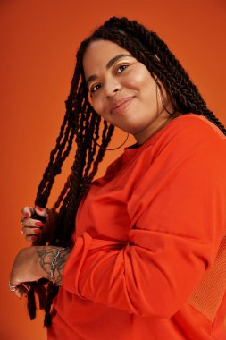 Örgülü bir kadın parlak turuncu bir arka plana karşı canlı bir elbiseyle gülümsüyor..