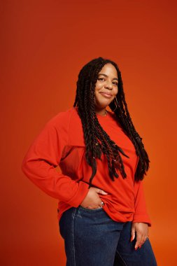 Örgülü kendine güvenen bir kadın şık bir kıyafet içinde turuncu bir fona gülümsüyor..