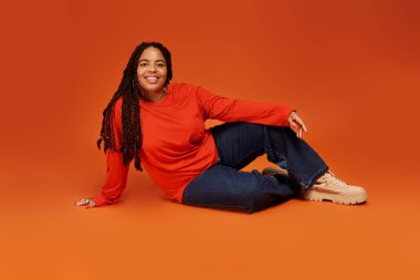 Güzel örgülü bir kadın turuncu arka planda şık bir elbiseyle rahatça poz verir..