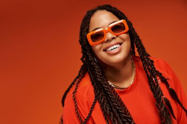 Saçları örgülü neşeli siyah bir kadın turuncu bir arka planda şık kıyafetini ve güneş gözlüğünü gösteriyor..