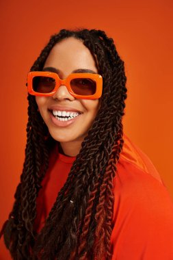 Saçları örgülü neşeli bir kadın turuncu bir fona karşı parlak turuncu bir elbiseyle poz veriyor..