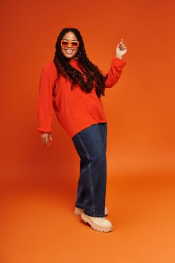Parlak giyimli Afro-Amerikan kadın cesur bir turuncu arka plana karşı özgüvenle poz veriyor.