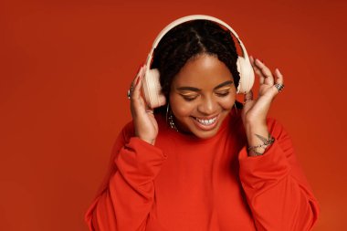 Canlı giyinmiş, örgülü neşeli kadın turuncu arka planda müziğin keyfini çıkarırken gülümsüyor..