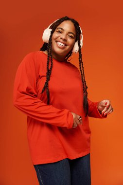 Örgülü neşeli bir Afrikalı Amerikalı kadın, kulaklıklı, parlak bir kıyafet içinde poz veriyor.