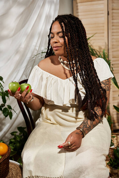 A beautiful Black woman with braids poses with fresh apples amid lush plants.