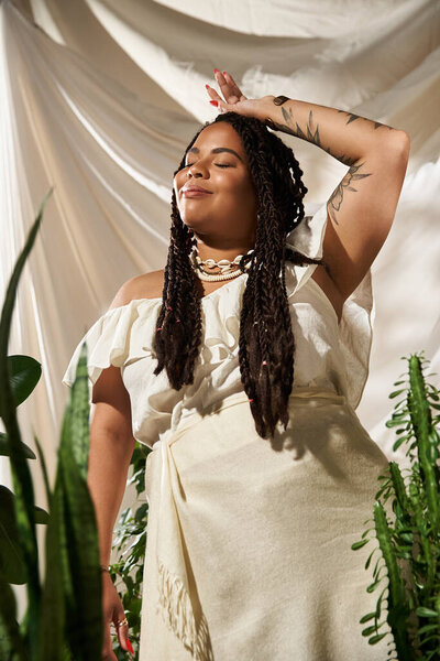 A pretty African American woman with braids poses gracefully among lush green plants.