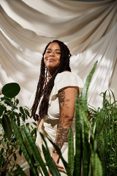 A beautiful African American woman with braids poses among lush green plants in the studio.
