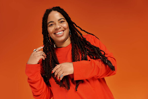 A confident Black woman in a bright outfit beams joy against a vivid orange backdrop.