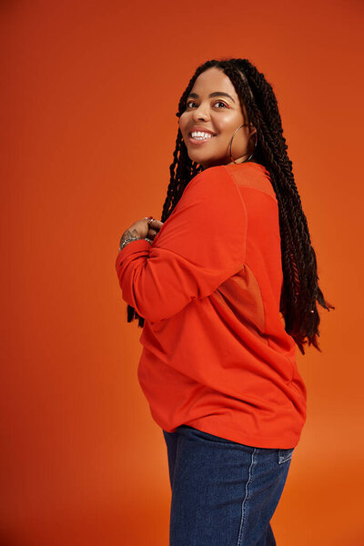 Стильная женщина с косичками позирует на ярком оранжевом фоне, выставляя напоказ свою моду.