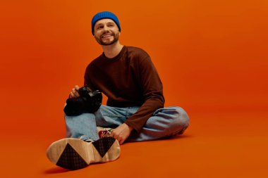 Neşeli genç bir adam yerde oturuyor, elinde parlak turuncu arka planı olan bir kamera var..