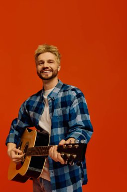 Gülümseyen genç adam mavi kareli gömlek giyerken akustik gitarını tıngırdatıyor..