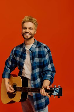 Gitarlı genç adam cesur bir turuncu arka planda dururken parlak bir şekilde gülümsüyor..