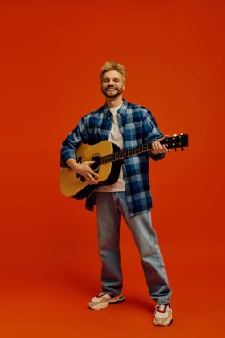 Gitar tıngırdatan neşeli genç bir adam, sıradan giyinmiş, çarpıcı turuncu bir arka planı var..
