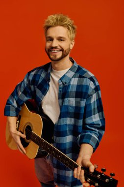 Büyüleyici genç adam gitarını neşeli bir şekilde tıngırdatıyor, canlı turuncu bir arka planla çevrili..
