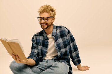 Yakışıklı genç adam yerde rahatça oturuyor, gülümserken kitap okumaya dalmış..