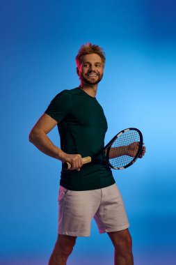 Yakışıklı genç adam sanatsal bir ortamda tenis raketini tutarken özgüvenle gülümsüyor..