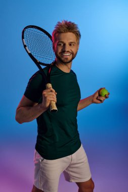 Genç adam renkli bir ortamda tenis raketi ve top tutarken dostça bir gülümseme sergiliyor..