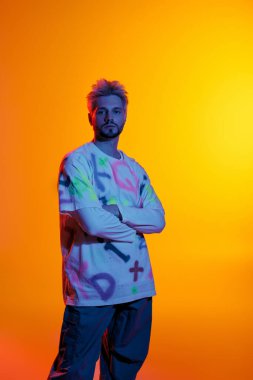 Yakışıklı genç bir adam kollarını kavuşturup eşsiz tarzını parlak bir ortamda sergiliyor..