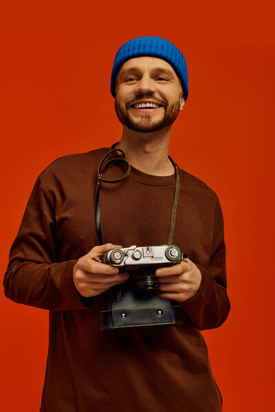 Handsome young man smiles widely while holding a classic camera in front of a bold orange wall.