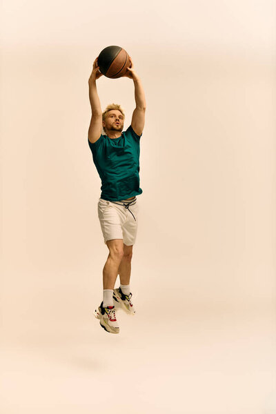 A handsome young man leaps high with a basketball, demonstrating athleticism in a studio setting.