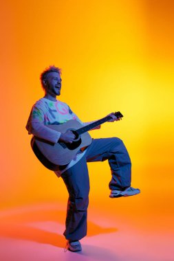 Kendine güvenen genç bir adam gitarını parlak renklerle çevrili bir şekilde tek ayak üstünde dans ederken tıngırdatır..