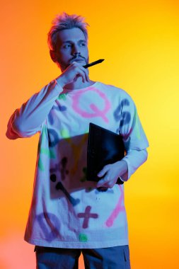 Yakışıklı genç bir adam elinde bir defter ve kalemle parlak turuncu bir zemin üzerinde duruyor..