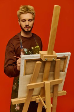 Çarpıcı özellikleri olan genç bir adam, parlak turuncu arkaplanlı bir sehpaya tutkuyla resim çizer..