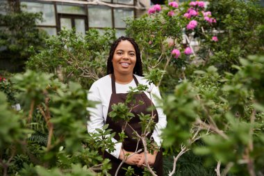Saçları örgülü gülümseyen Afrikalı Amerikalı bir kadın canlı yeşillikler ve çiçekler arasında duruyor..