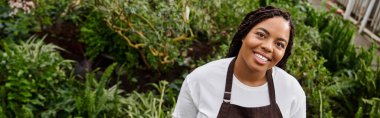 Saçları örgülü Afrikalı bir Amerikalı kadın yemyeşil bir serada bitkilerle ilgilenir. Mutluluk yayar, afiş yapar.
