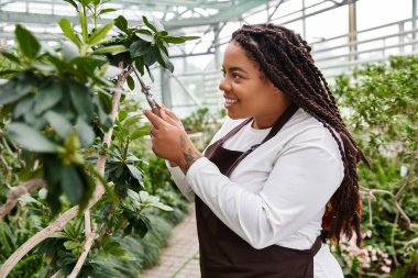 Saçları budanmış, gülen siyah bir kadın yemyeşil bir serada bitkiler yetiştiriyor..