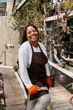 Saçları örgülü gülümseyen Afro-Amerikan bir kadın bir serada dikiliyor, yemyeşil bitkilerle ilgileniyor..