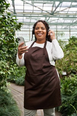 Canlı bir serada bitkilerle ilgilenirken örgü ören Afrikalı bir Amerikalı kadın gülümsüyor..