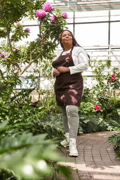 Saçları örgülü bir Afrikalı Amerikalı kadın çiçeklerle dolu canlı bir serada bitki yetiştiriyor..