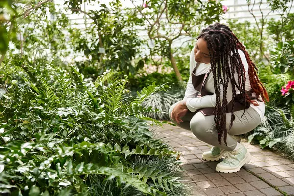 Saçları örülü siyah bir kadın güneşli bir serada canlı bitkilerle ilgilenir..