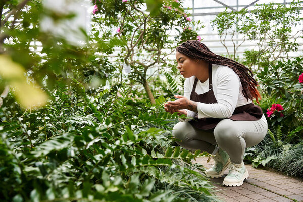 An African American woman tending to lush plants in a greenhouse, enjoying her gardening time.