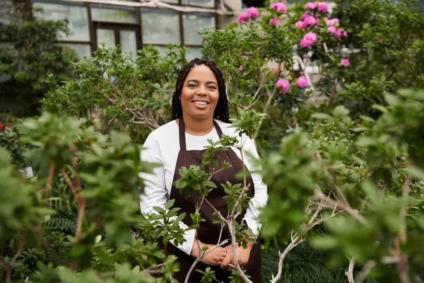 Saçları örgülü gülümseyen Afrikalı Amerikalı bir kadın canlı yeşillikler ve çiçekler arasında duruyor..