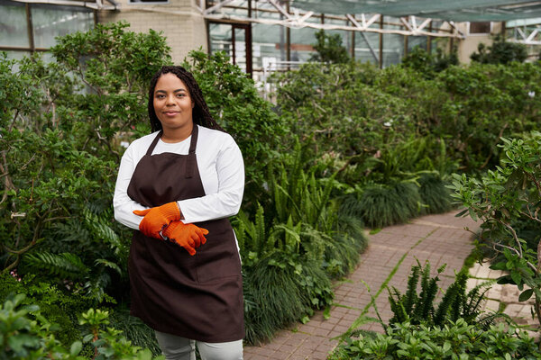 African American woman with braids enjoys her time gardening amidst lush greenery in a greenhouse.