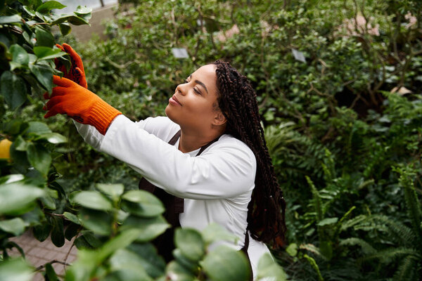 An African American woman tends to plants in a lush greenhouse, showcasing her gardening skills.