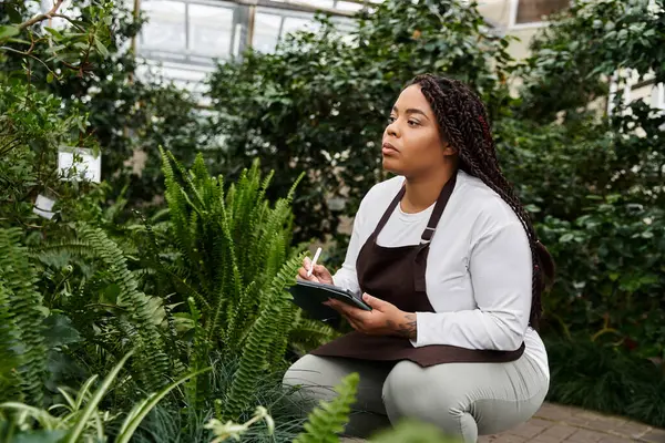 Örgülü Afrikalı bir Amerikalı kadın çiçeklerle ilgileniyor, sera vahasında notlar alıyor..