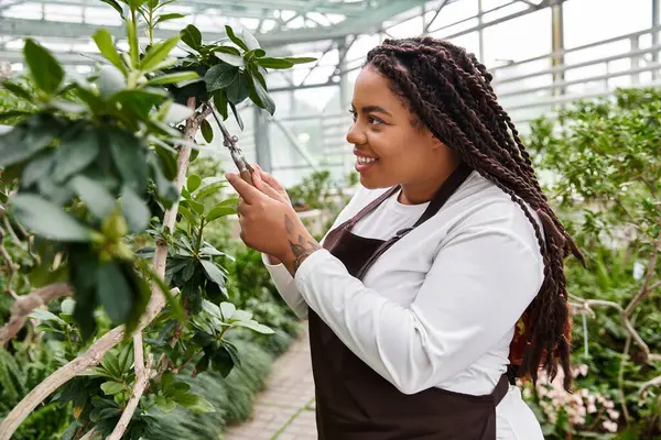 Saçları budanmış, gülen siyah bir kadın yemyeşil bir serada bitkiler yetiştiriyor..