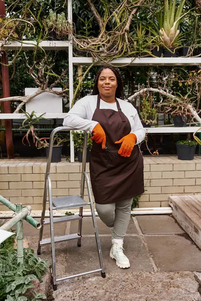 Afro-Amerikalı bir kadın bir serada güvenle duruyor, yeşillik ve bitki yetiştiriyor..