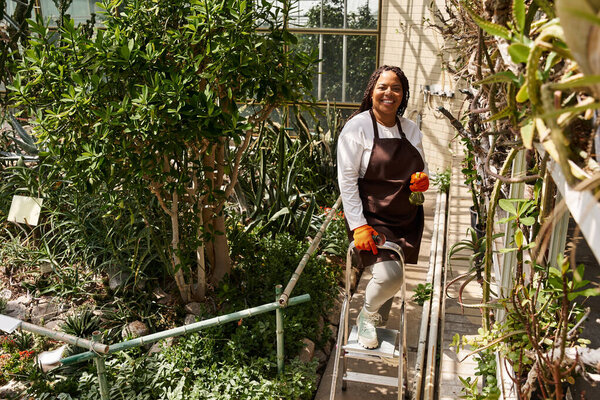 A joyful Black woman with braids tends plants in a vibrant greenhouse full of greenery.