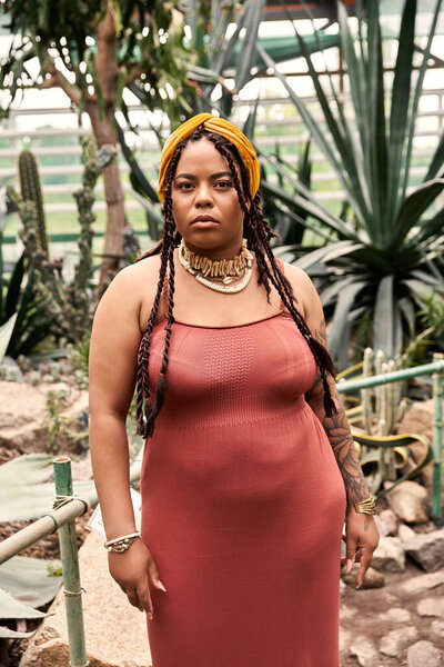 A stylish African American woman with braids stands confidently amid beautiful plants indoors.