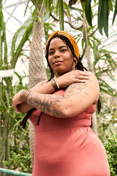 A pretty African American woman with braids poses gracefully among lush plants in a greenhouse.