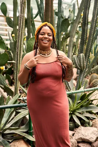 Saçları örgülü gülümseyen Afrikalı bir Amerikalı kadın bir serada yemyeşil bir alanla çevrili..